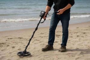 Henkilö käyttää metallinpaljastinta hiekkarannalla meren äärellä. Laite on lähellä maanpintaa ja sitä liikutetaan rauhallisesti edestakaisin rantahiekalla.